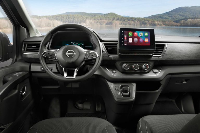 Abitacolo di un furgone Nissan con volante logato, display touch con app, comandi al cruscotto e vista su un lago